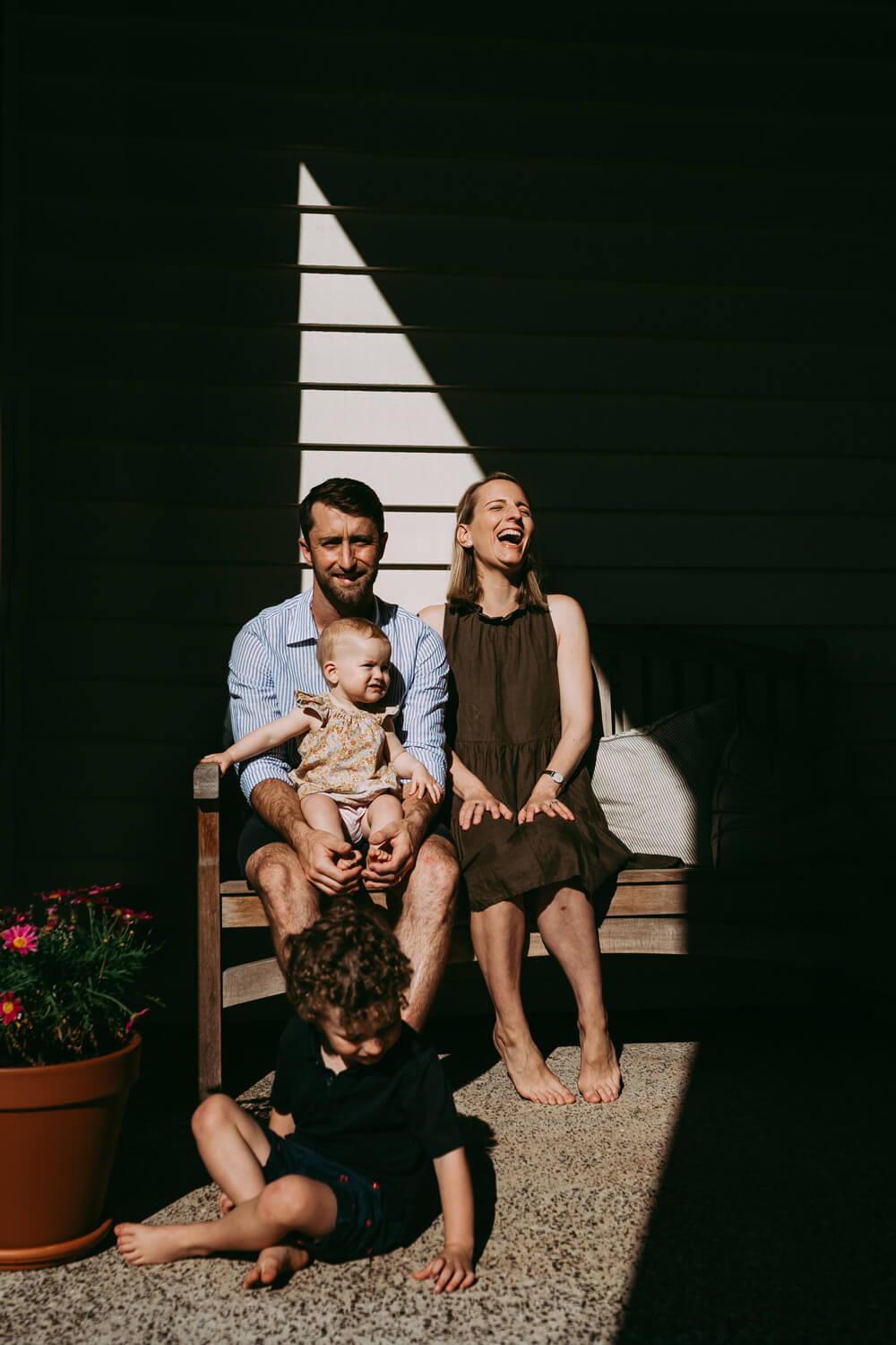 A family of four sits on a wooden bench. A woman is laughing as she throws her head back. They sit in a triangle of light, dark shadow surrounds them.