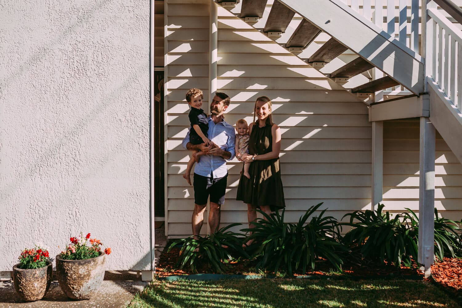 A family of four stands under a set of stairs. Light shines through the stairs and casts stripes across the family.