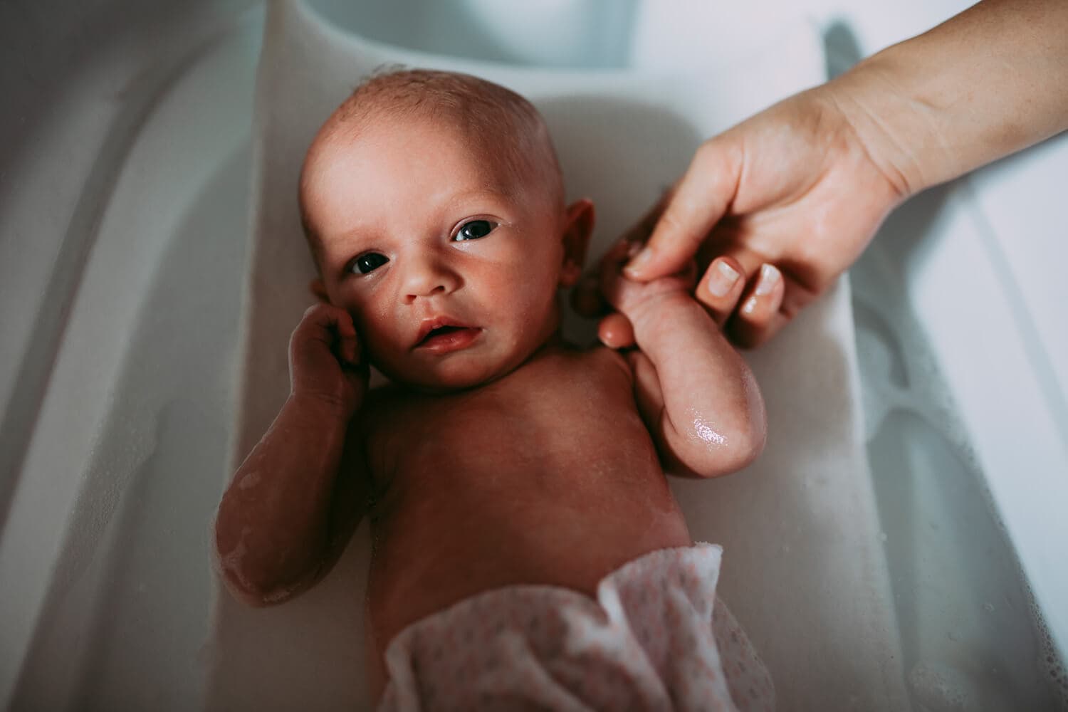 A newborn baby hold its mothers hand as it lies in a bath.