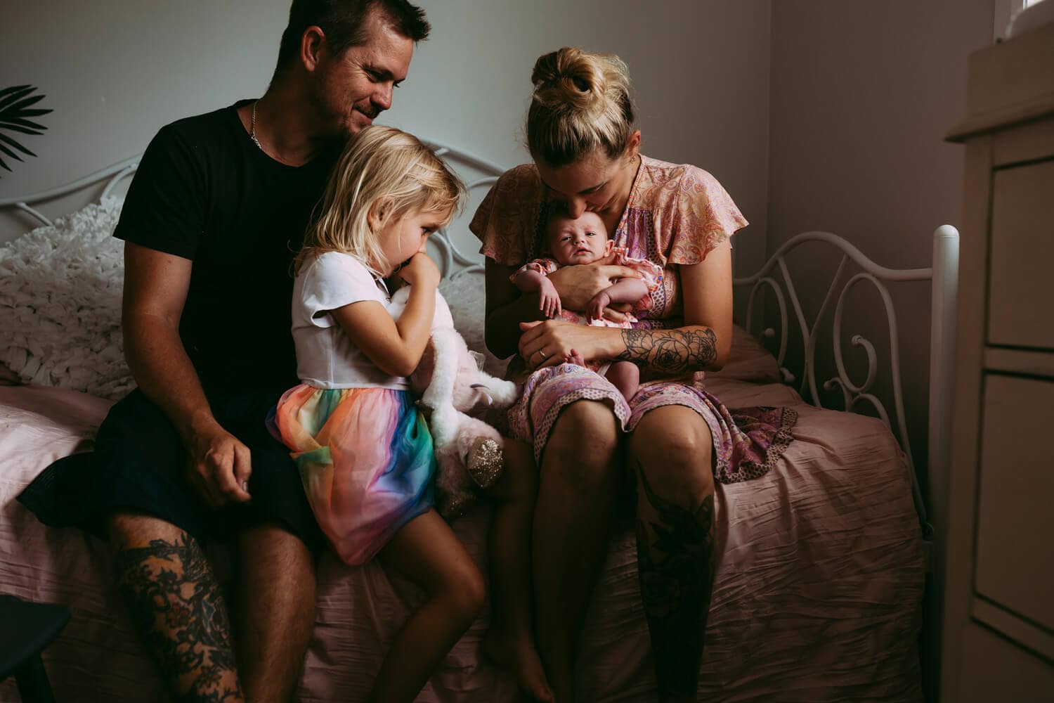 A family of four sit on a bed, staring down at their newborn.