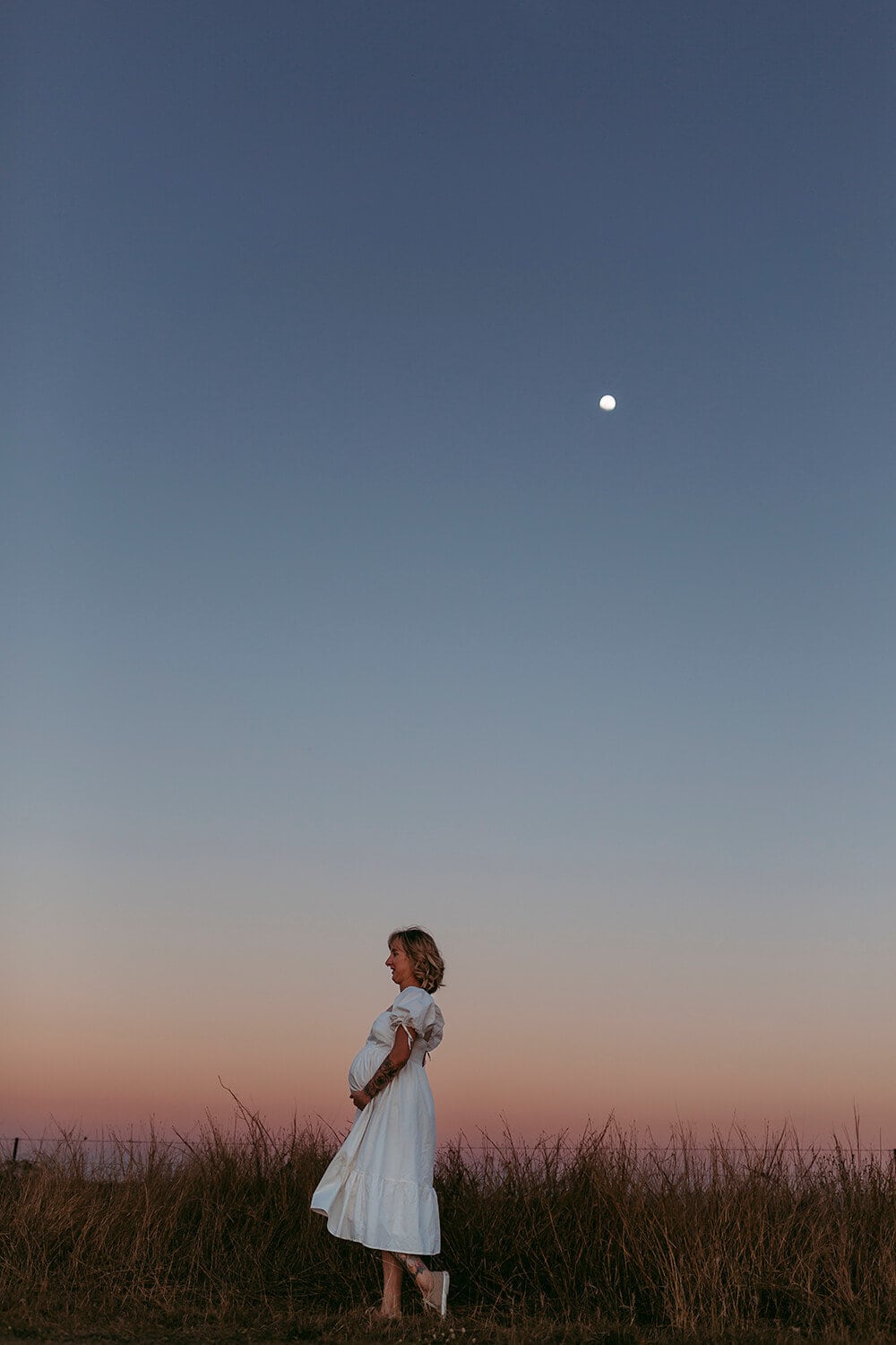 A pregnant woman wearing a white dress holds her belly. The moon shines above her.