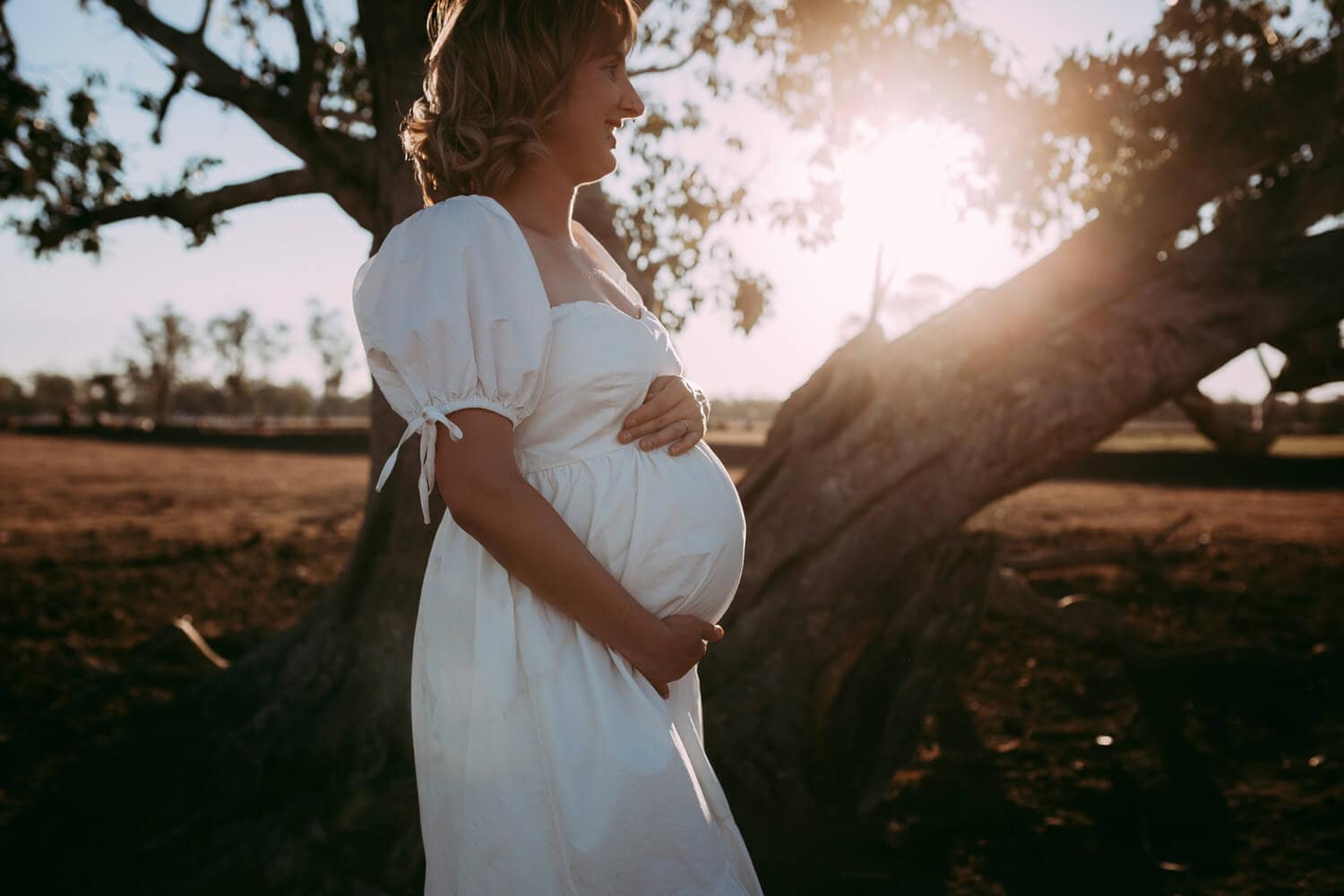 A pregnant woman wearing a white dress places her hand around her belly. A warm sun beams through the tree behind her.