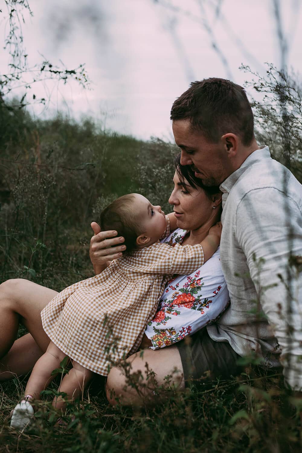 A toddler wraps her arms around her mother's neck as her father watches on. They're all sitting in a grassy field.