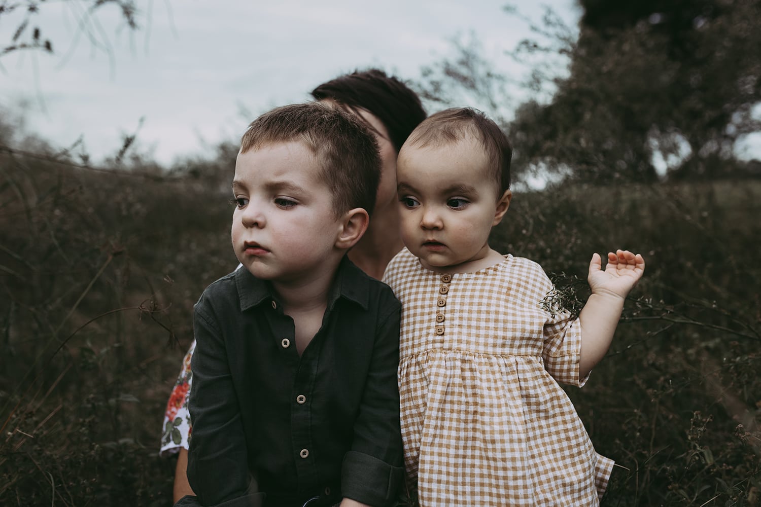 A little boy and girl stand next to one another. Both are snuggled close with a serious face. Their mother sits behind them.