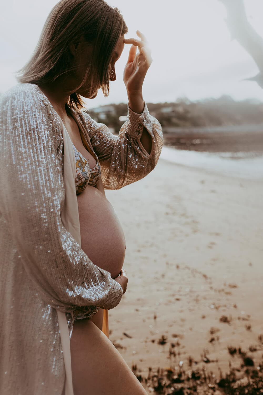 A woman brushes hair from her face and gently holds her other hand on her pregnant belly. She's at the beach.