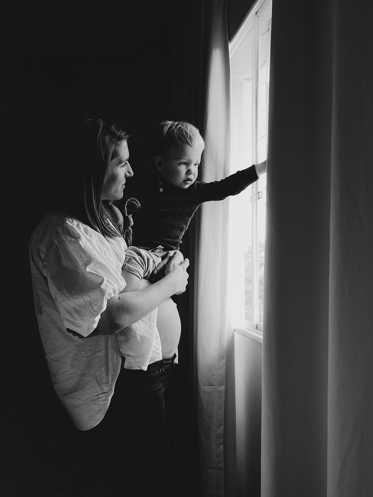 A pregnant mother holds her toddler on her belly as they look out a window.