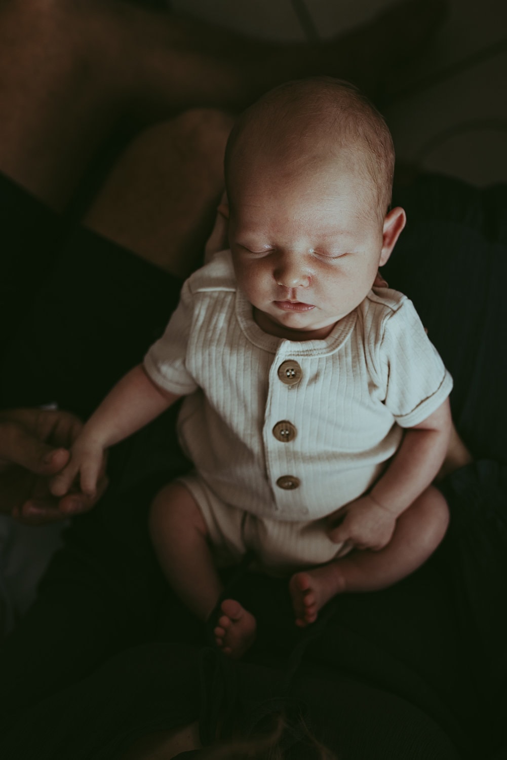 A newborn baby is asleep while being held upright by her mother. Shadow surrounds the baby.