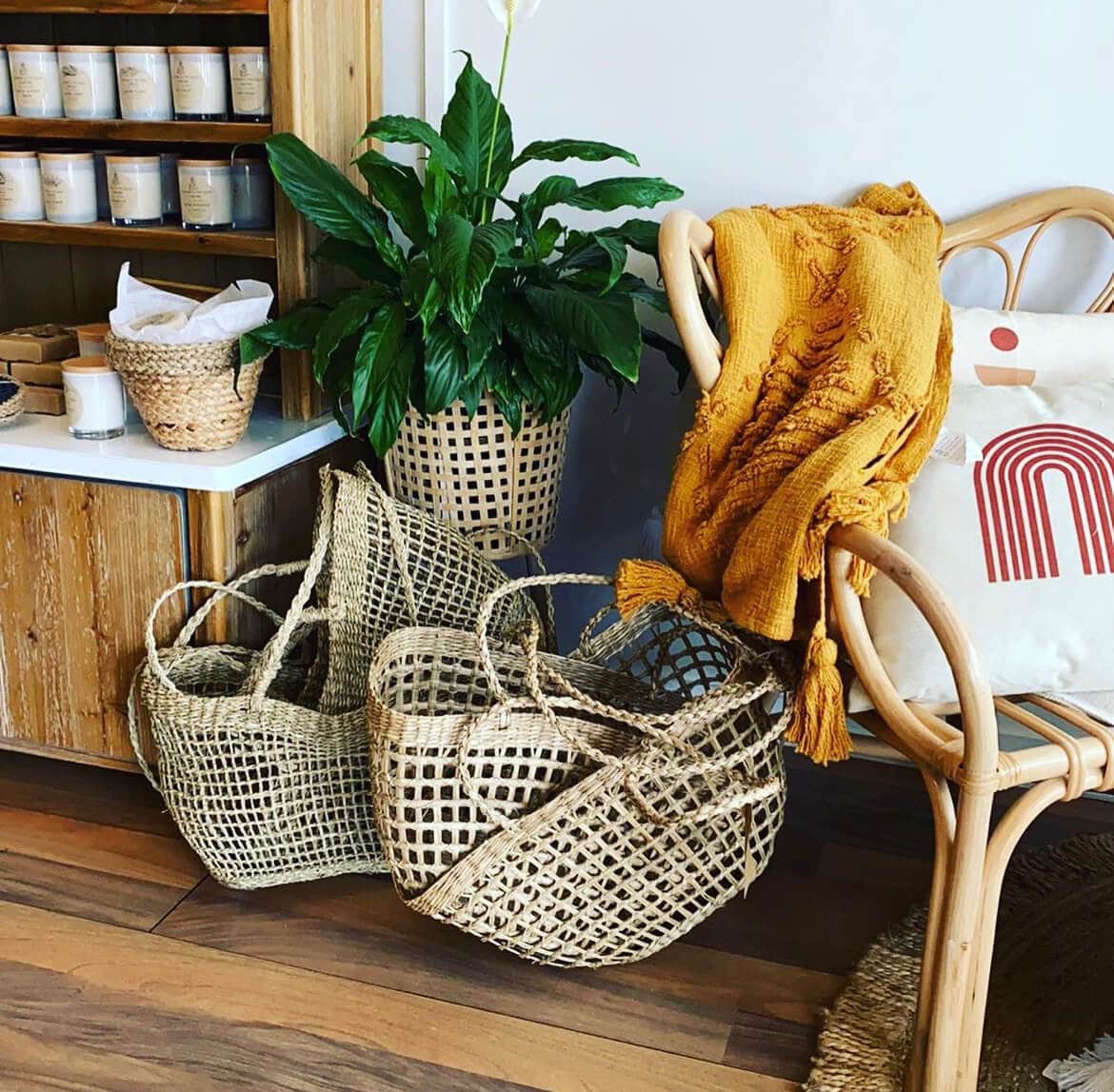 A can chair with a cushion on it and a mustard coloured throw hangs off it. A group of baskets sits beneath it. Soy Candles sit on a shelf behind it.