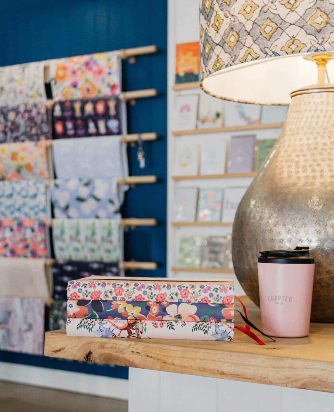 A collection of three floral notebooks sits on a table with a lit lamp. Reams of wrapping paper and cards can be seen hanging on the wall in the background.