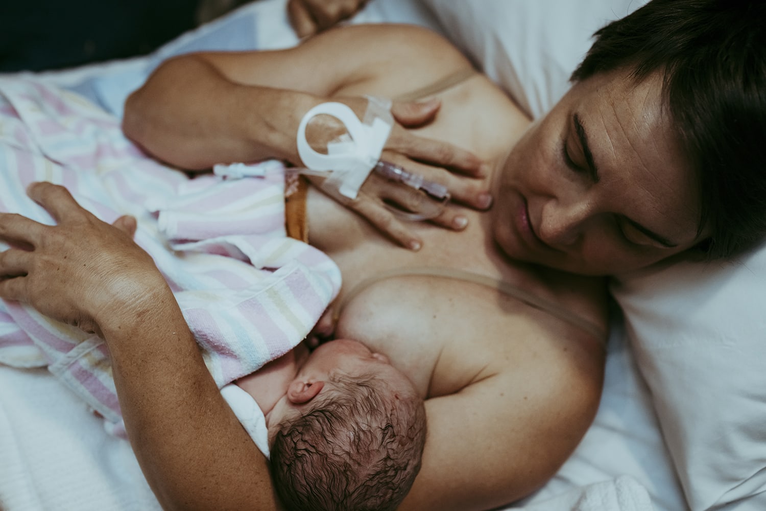 A mother has a tube connected to her hand, as she lies back in a hospital bed, cradling her newborn as they breastfeed.