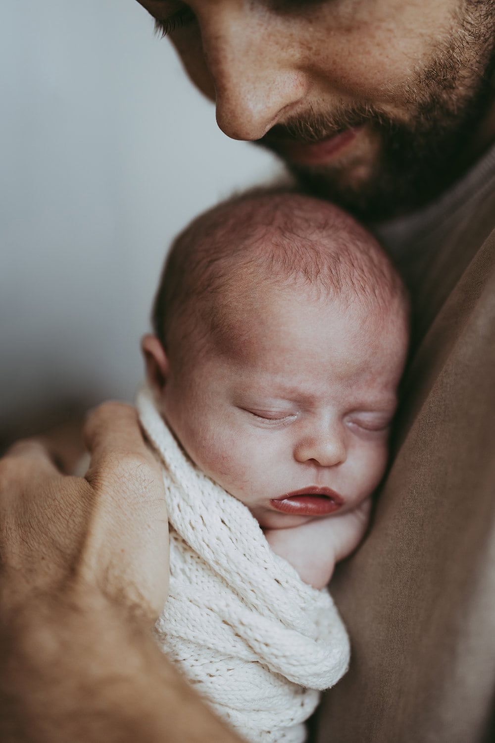 A father holds his newborn baby against his chest. The baby is wrapped snuggly in a cream blanket and has her hand resting under her chin. 