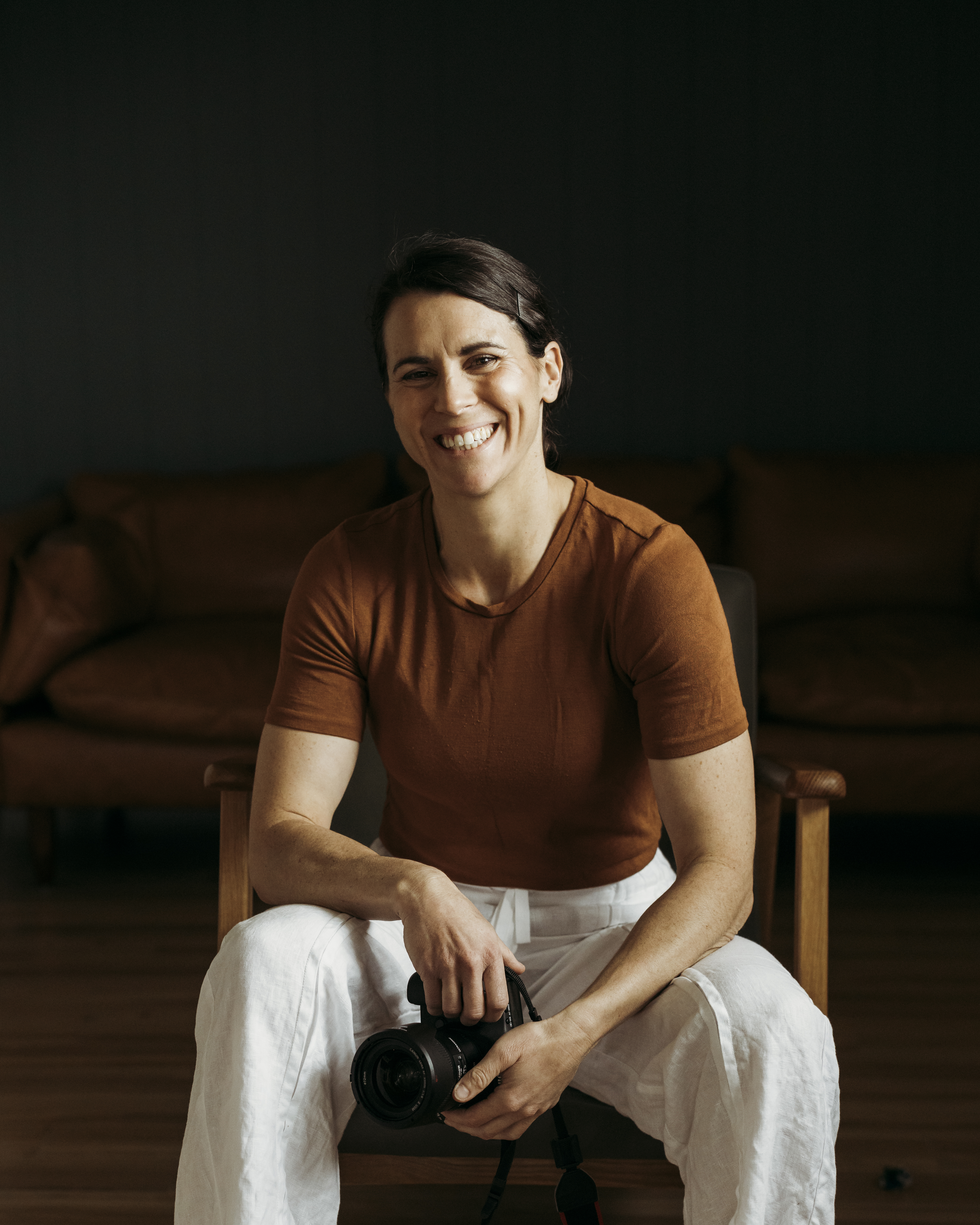 A woman sits on a chair looking at the camera smiling. She wears a warm brown structured t-shirt and white linen pants. She casually rests both arms on her legs as she holds a camera.