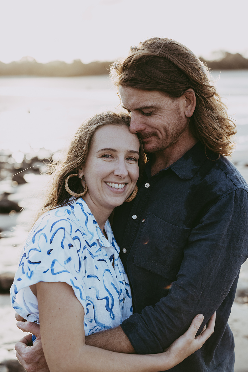 A couple hold one another closely as the sun sets in the background. They are at the beach. 