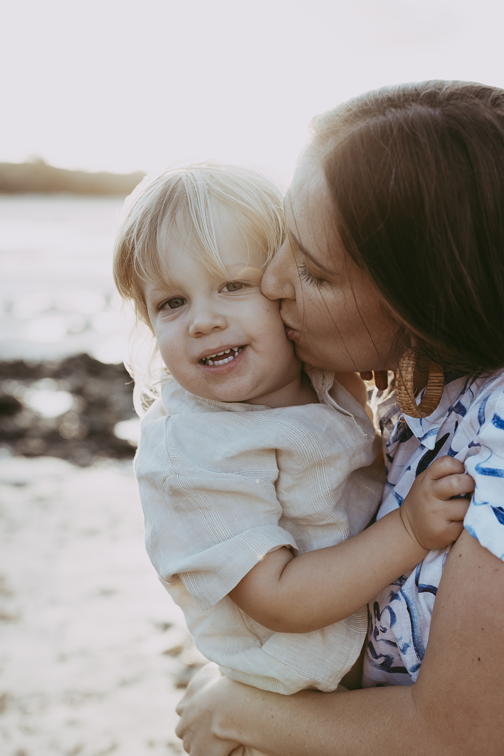 A mother squeezes her toddler tight as she kisses his cheek. The sun illuminates them from behind. 