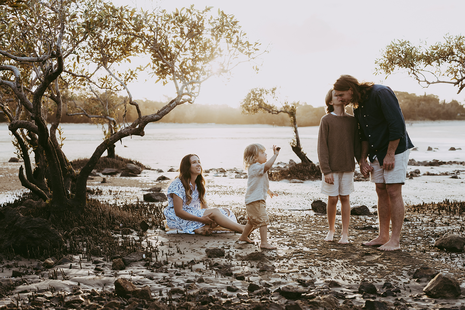 A family of four is exploring a beach. They are framed by mangroves as the sun sets behind them.