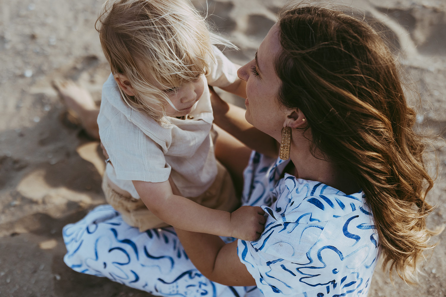 A mother and child embrace. The view is from above them as they sit in sand.