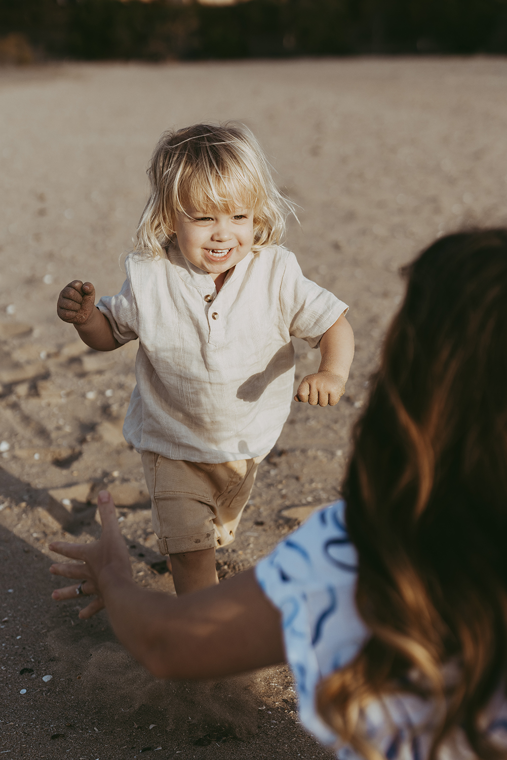 A toddler runs towards his mothers arms, which are outstretched in the corner of the photograph. He's smiling at her.