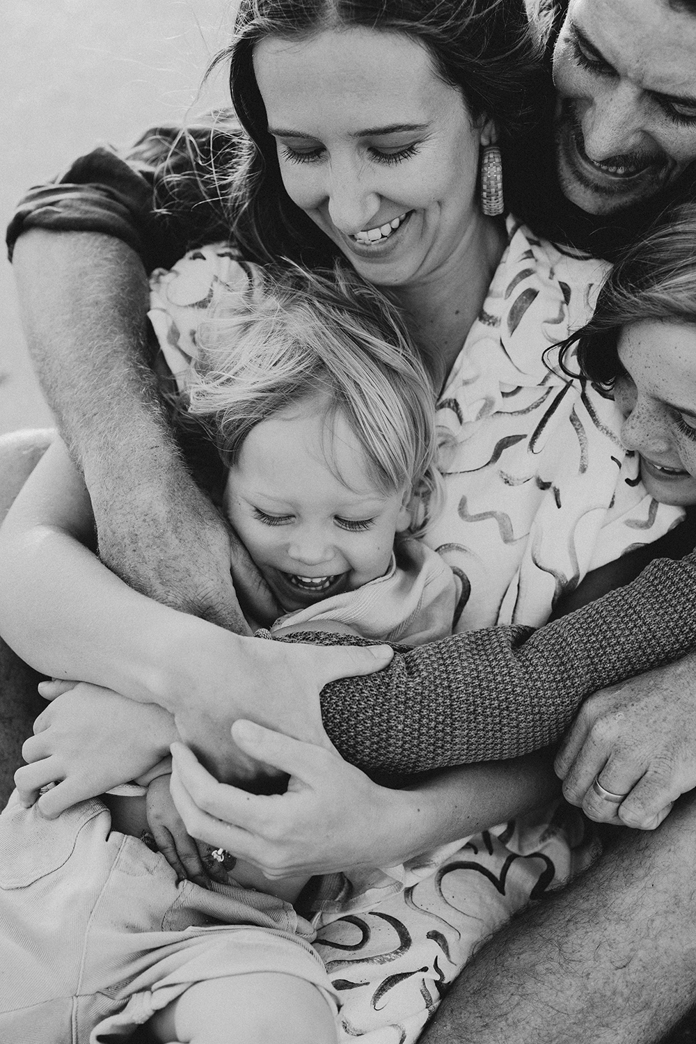 A family of four cuddle each other tightly as the youngest child giggles. All eyes are on him. The image is in black and white. 