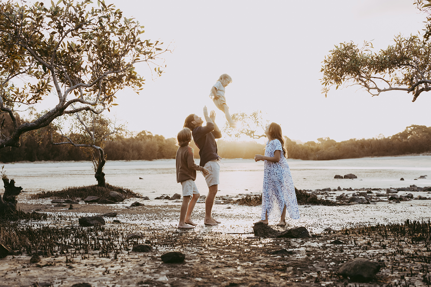 A family of four plays on a beach. The father is throwing his laughing youngest up in the air while his brother and mother watch and smile at the action. They are framed my mangroves as the sun sets.