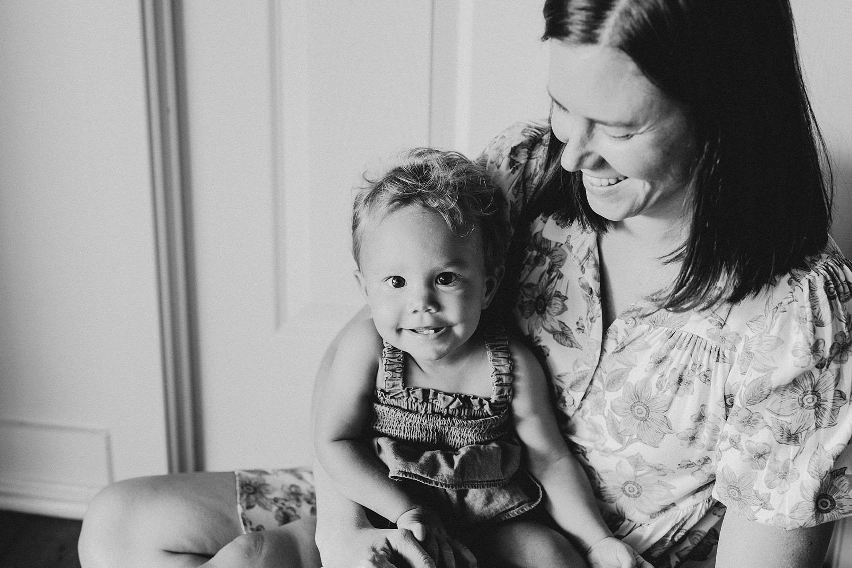 A black and white image of a smiling toddler sitting on her mother's lap. Her mother is looking down at her smiling.