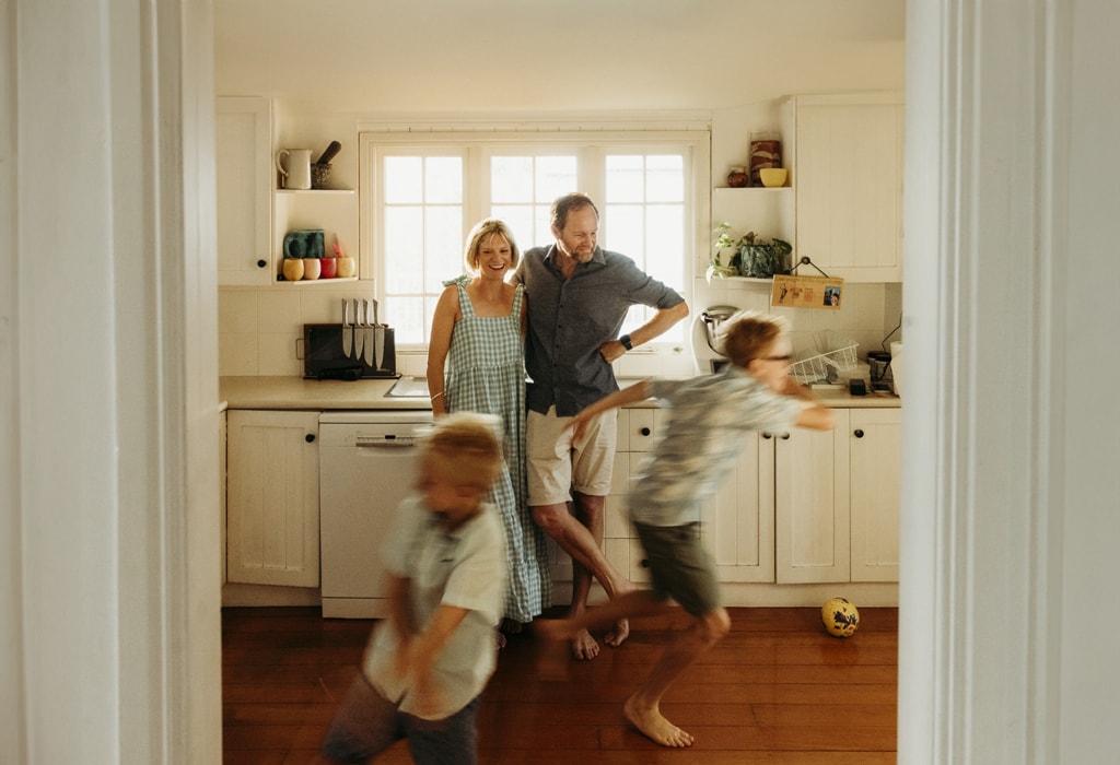 A couple stand in their kitchen with the arms around each, illuminated by the window behind them. Their two boys are blurred as they sprint past them. 