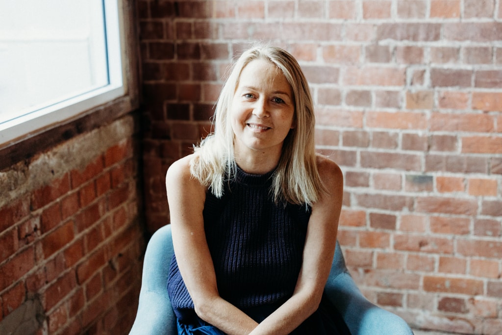 A woman smiles at the camera. She is sitting in a blue chair with her arms crossed over her lap. A red brick wall is in the background.