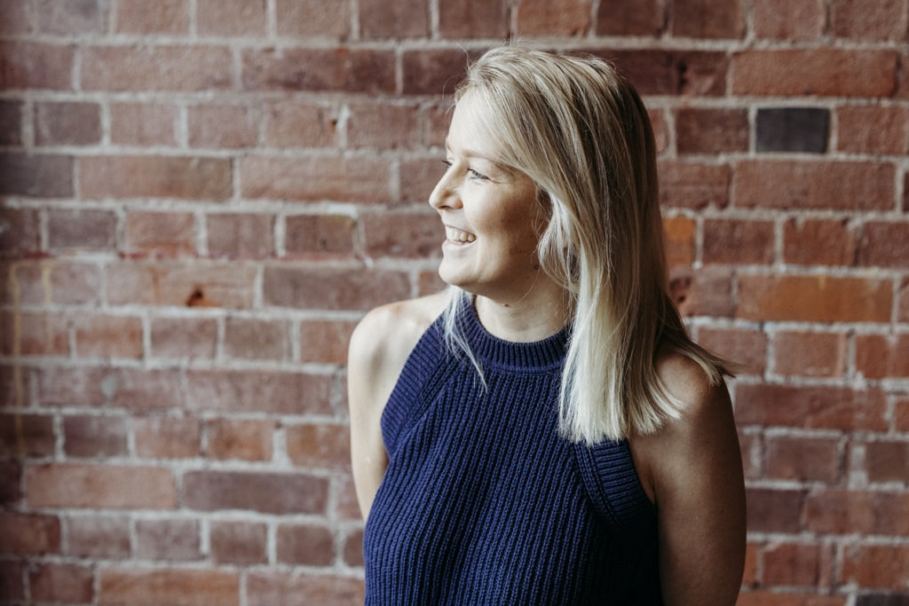 A woman smiles as she looks out of frame. She is standing in front of a red brick wall. She is Rockhampton naturopath Kate Willis.