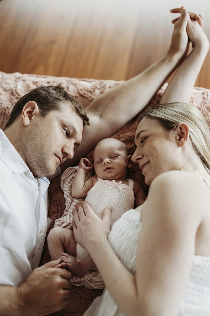 A newborn is asleep as her parents lie down next to her, both holding hands as they stare at her.