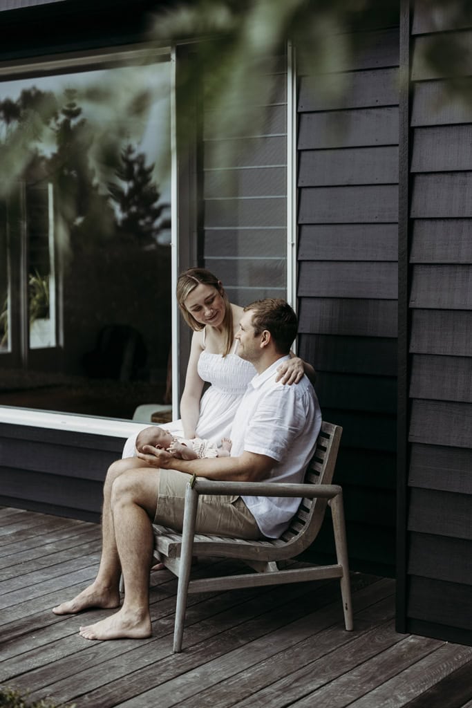 A couple sit on a wooden chair as they cradle their newborn.