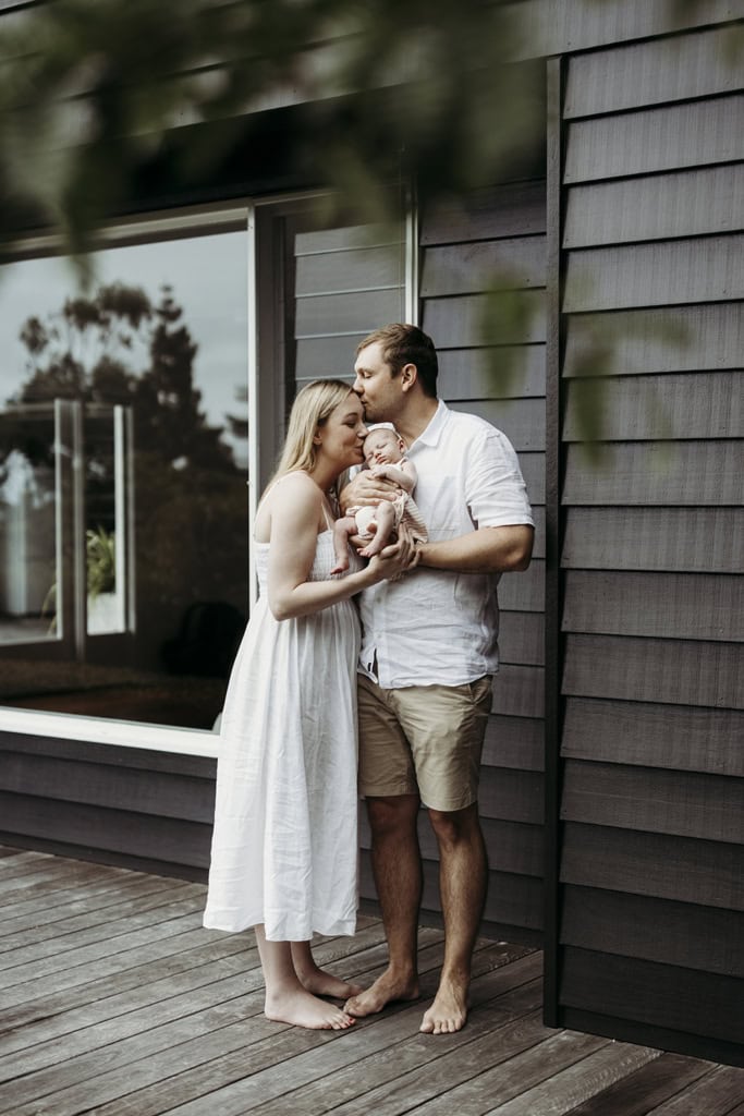 Parents cradle their newborn baby in their arms. The father kisses his wife on her head, while the mother kisses their newborn. They are standing on a grey timber deck. Black walls in the background, green leaves in the foreground frame them.