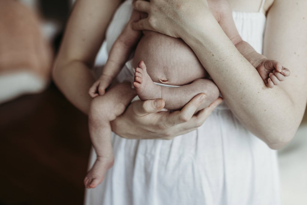 A mother cradles her newborn baby. The viewer can see the baby's belly button as one leg is curled up, almost against her belly. Her arms hand down by her side.