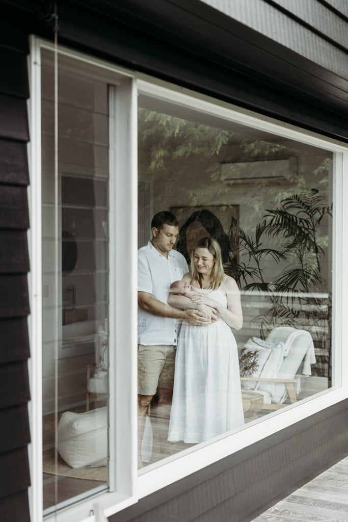 A couple hold their newborn. The image is looking through a window as the couple stare at their newborn.