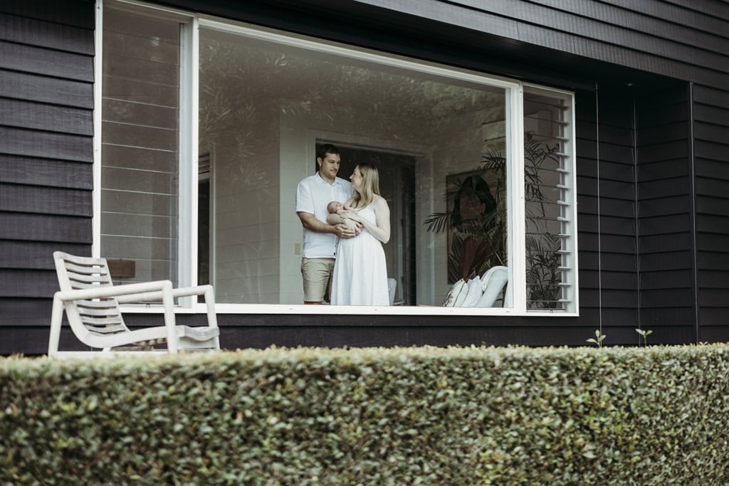 A man and woman hold their newborn baby and are framed by a window.