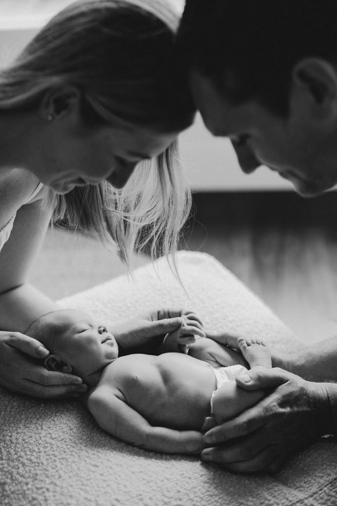 A mother and father have their heads together as they lean over their newborn baby. Their baby is curled up on a cushion. 