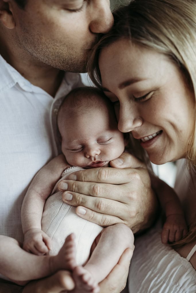 A man and woman cuddle their newborn baby. The father kisses the top of the mother's head as she snuggles with the baby.