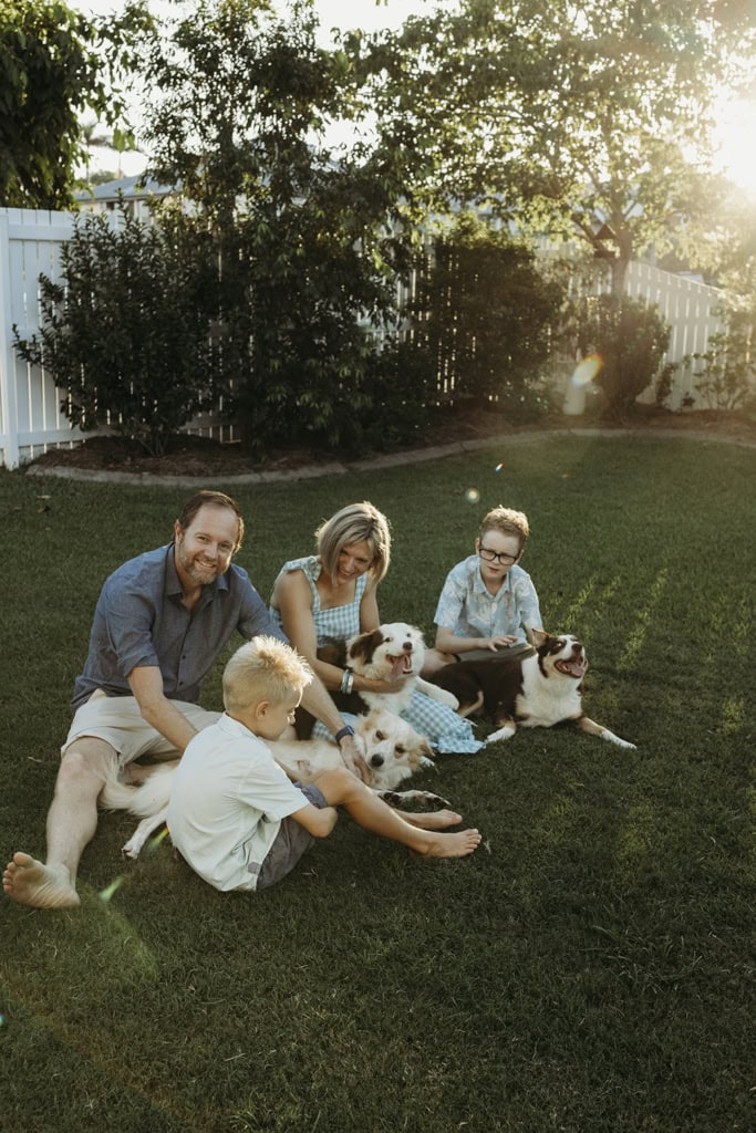 A family of four pat their three dogs while sitting in the backyard. The sun streaks through the trees behind them.