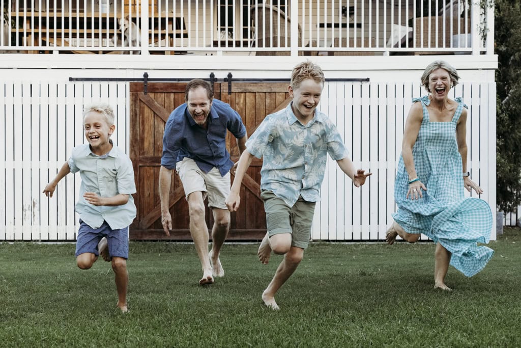 A family of four sprints across their lawn laughing. Dad is bending down slightly, chasing his two sons. All have joy on their face. Their white house is visible in the background.