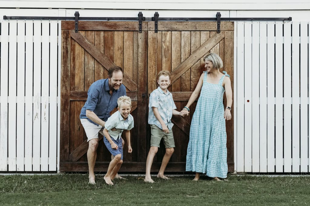 A family of four stand in front a big barn door. Dad is holding onto one of his sons, both smiling. Mum is holding her other son's hand and she is looking at him smiling. They are standing on grass.