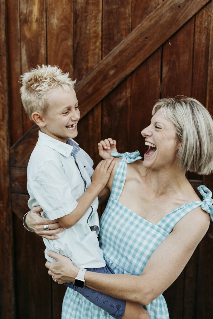 A young boy is held by his mother as they look at one another and laugh. Warmer timber is in the background.