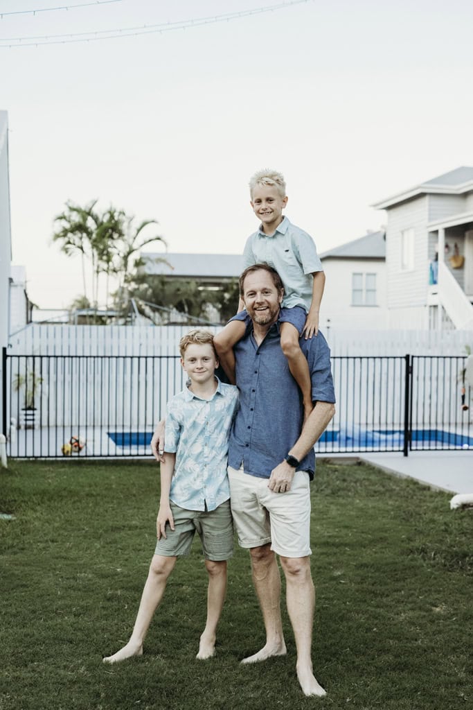 A father has one son on his shoulders and another next to him as they smile at the camera. A pool and while homes are visible in the background.