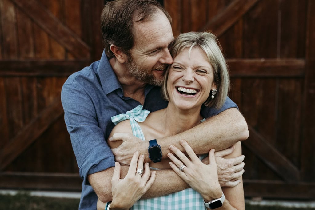 A man holds his wife shoulders as they embrace. He is snuggled into her and they are both laughing. Her hands are holding onto his arms.