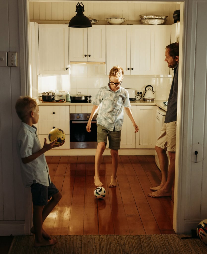 A young boy plays with a soccer ball in the family kitchen as his father looks on. His younger brother holds a ball and is pictured in the foreground, as he leans up against the doorway.