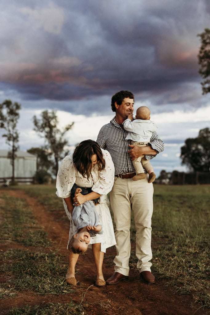 A mother holds her toddler son upside down, while a father stands next to them holding his infant son. They are standing in a red dirt paddock with small amounts of gras and an incredible stormy sky in the background.