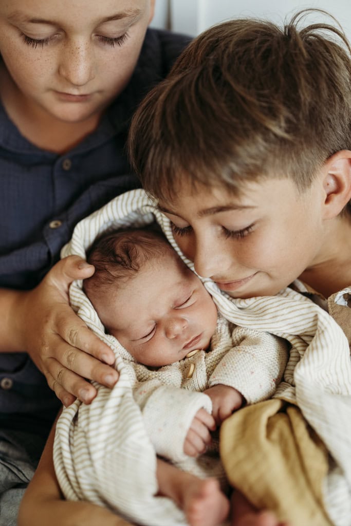 Two boys hold their baby brother as they both smile. 