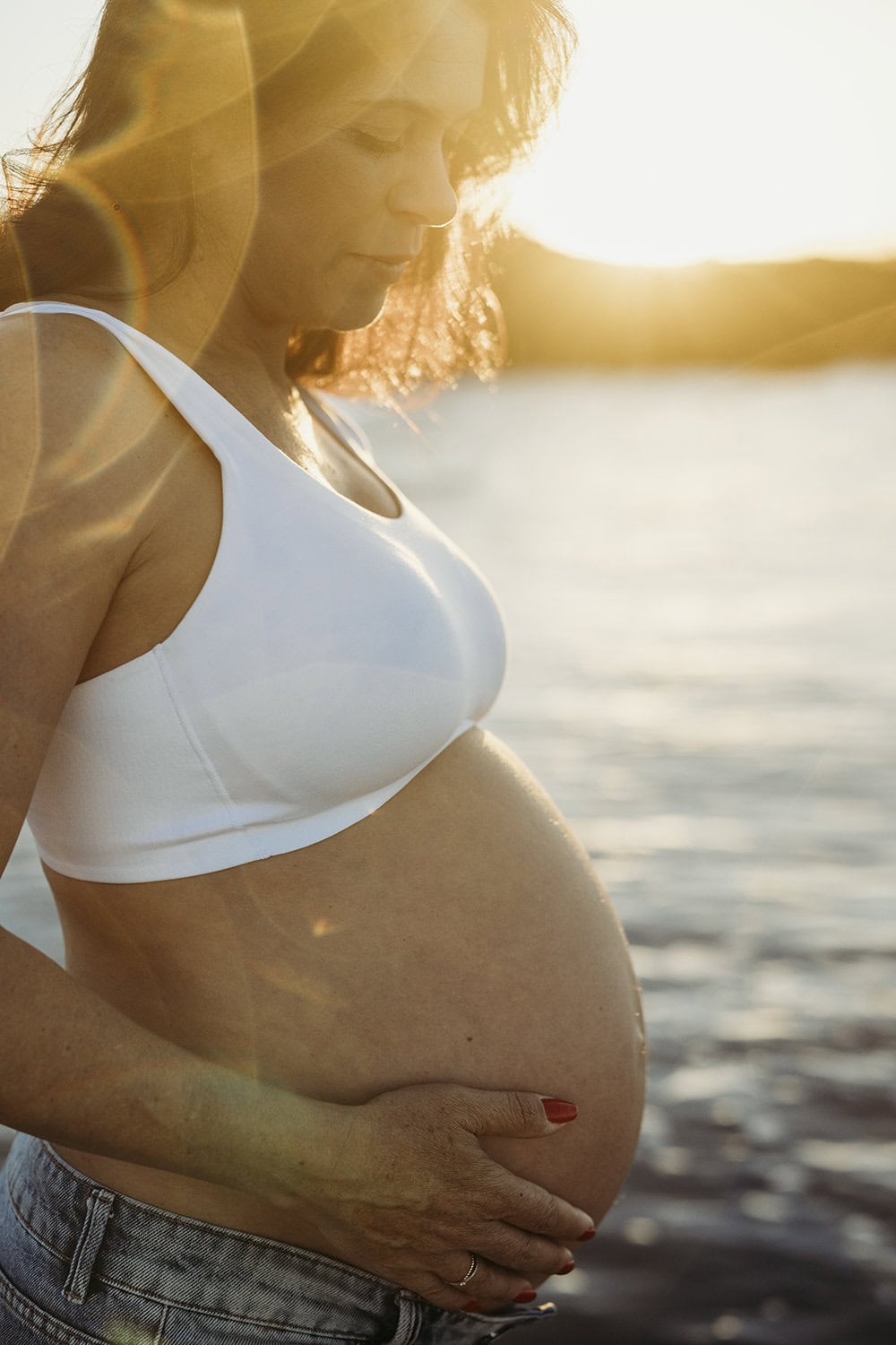 A woman looks down at her pregnant belly as the sun sets behind her at the beach.