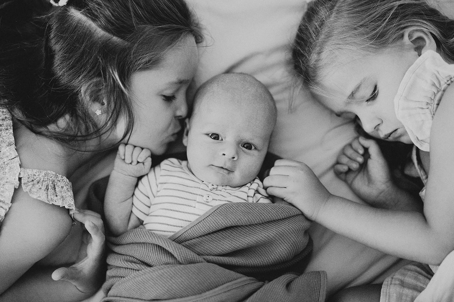 Two young girls lay next to their baby brother who is in between them. One girl is kissing his head, while the other holds his hand. 