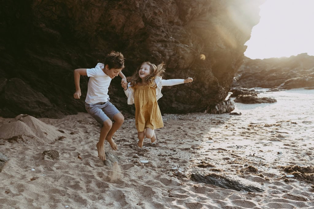 A boy and girl hold hands as they jump on a beach with a giant black rock behind them as the sun streams in. 