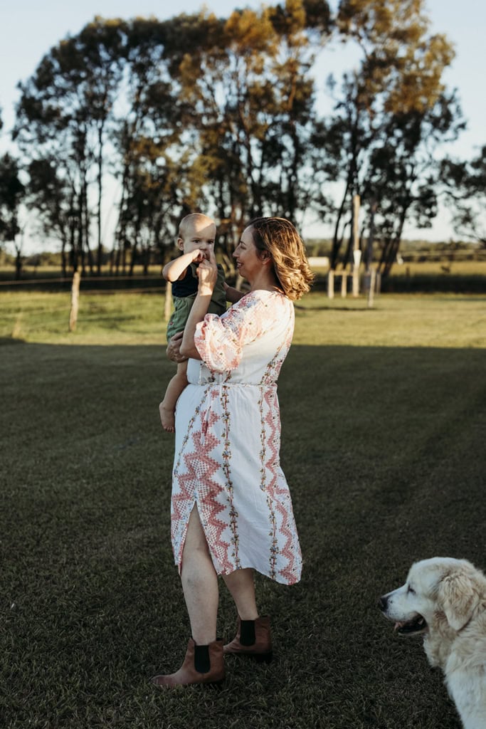 A toddler is held by his mother. She is smiling lovingly at him. They are standing outside with their dog next to them. 