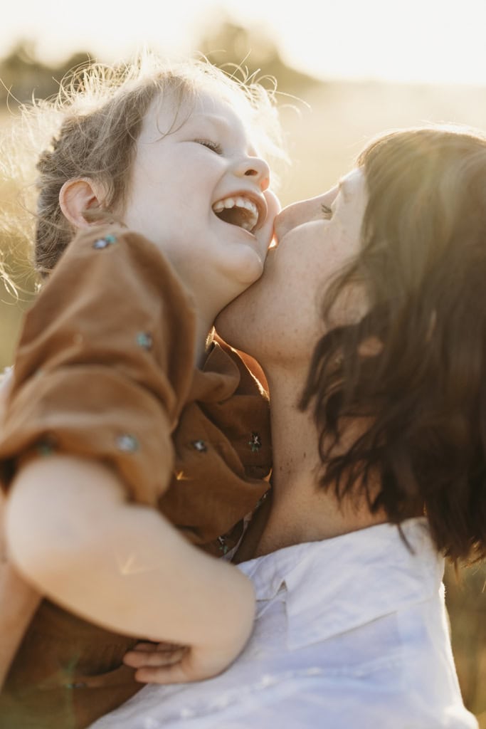 A mother kisses her daughter's cheek as she giggles. Sun shines from behind them. 