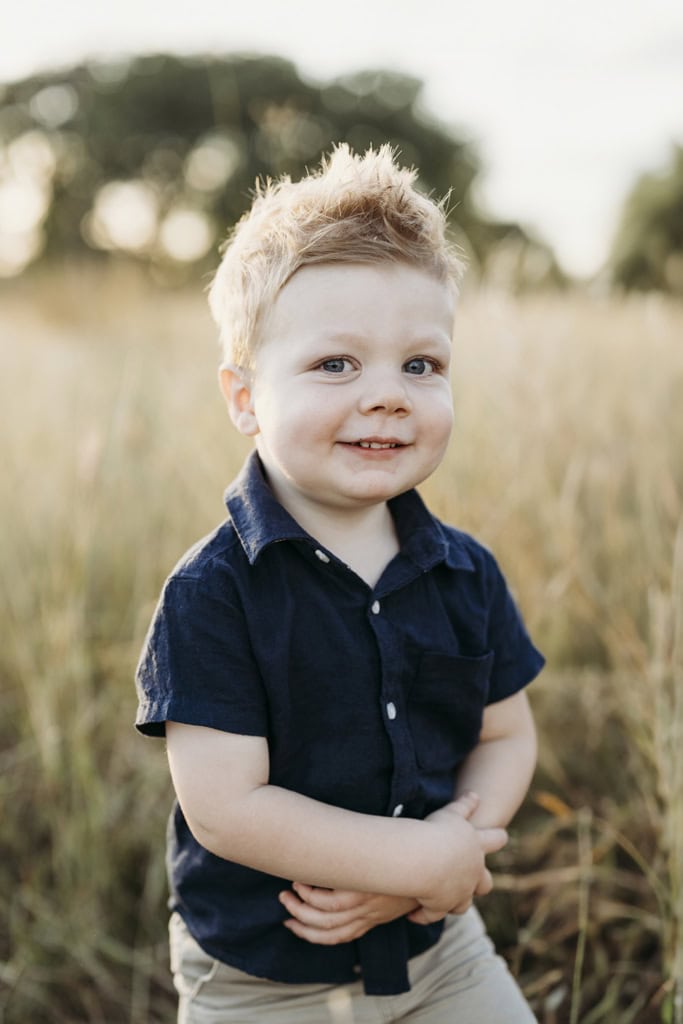 A little boy holds his hands across himself, smiling slightly at the camera. 